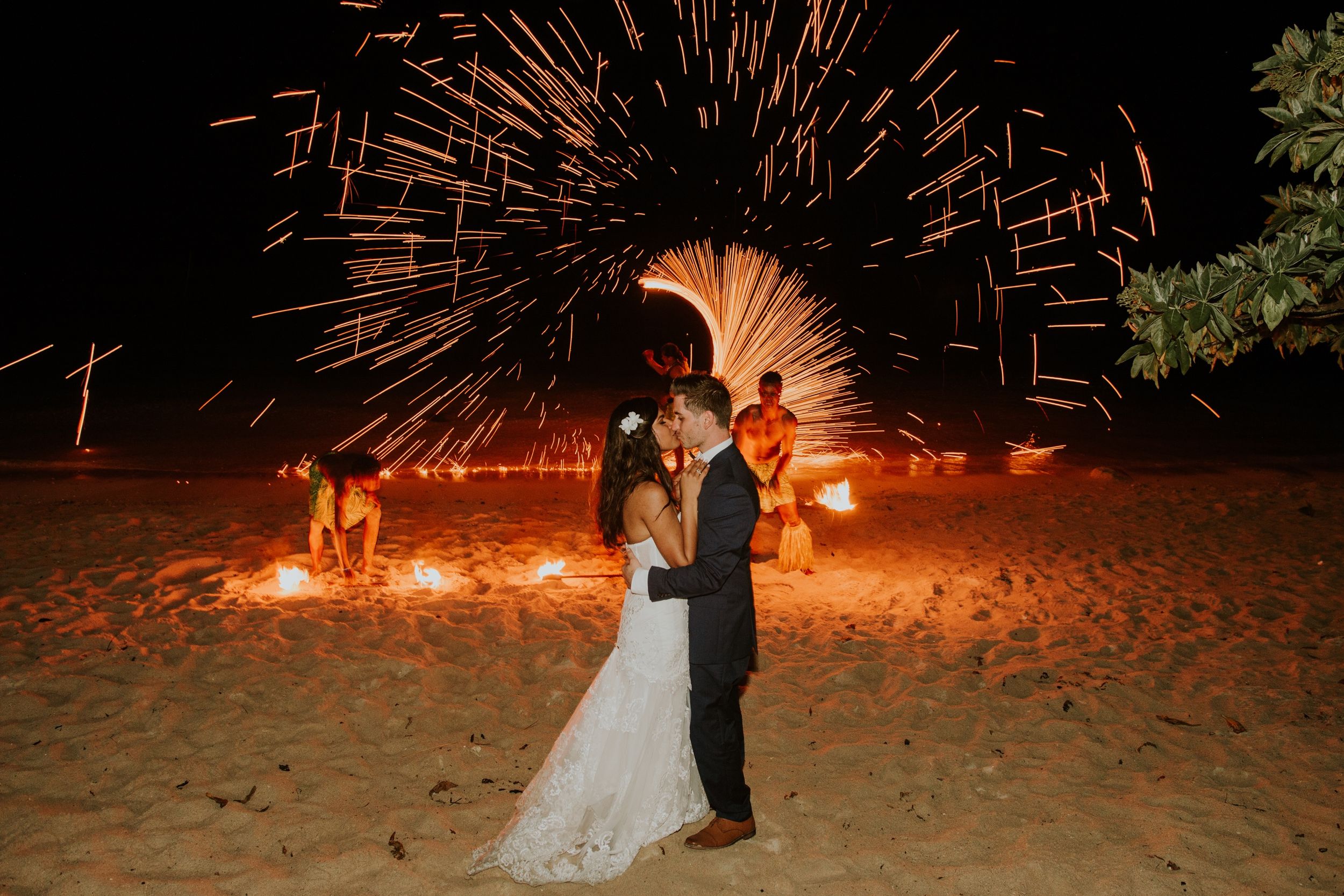 A romantic couple poses on a beach with spectacular fireworks display creating shimmering patterns behind them.
