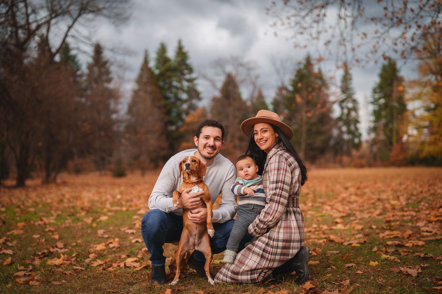 A family and their golden retriever pose together on fallen autumn leaves in a park setting.