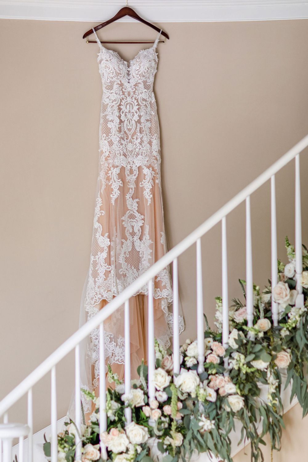 Elegant lace wedding gown hanging on wooden hanger against beige wall with floral staircase decoration.