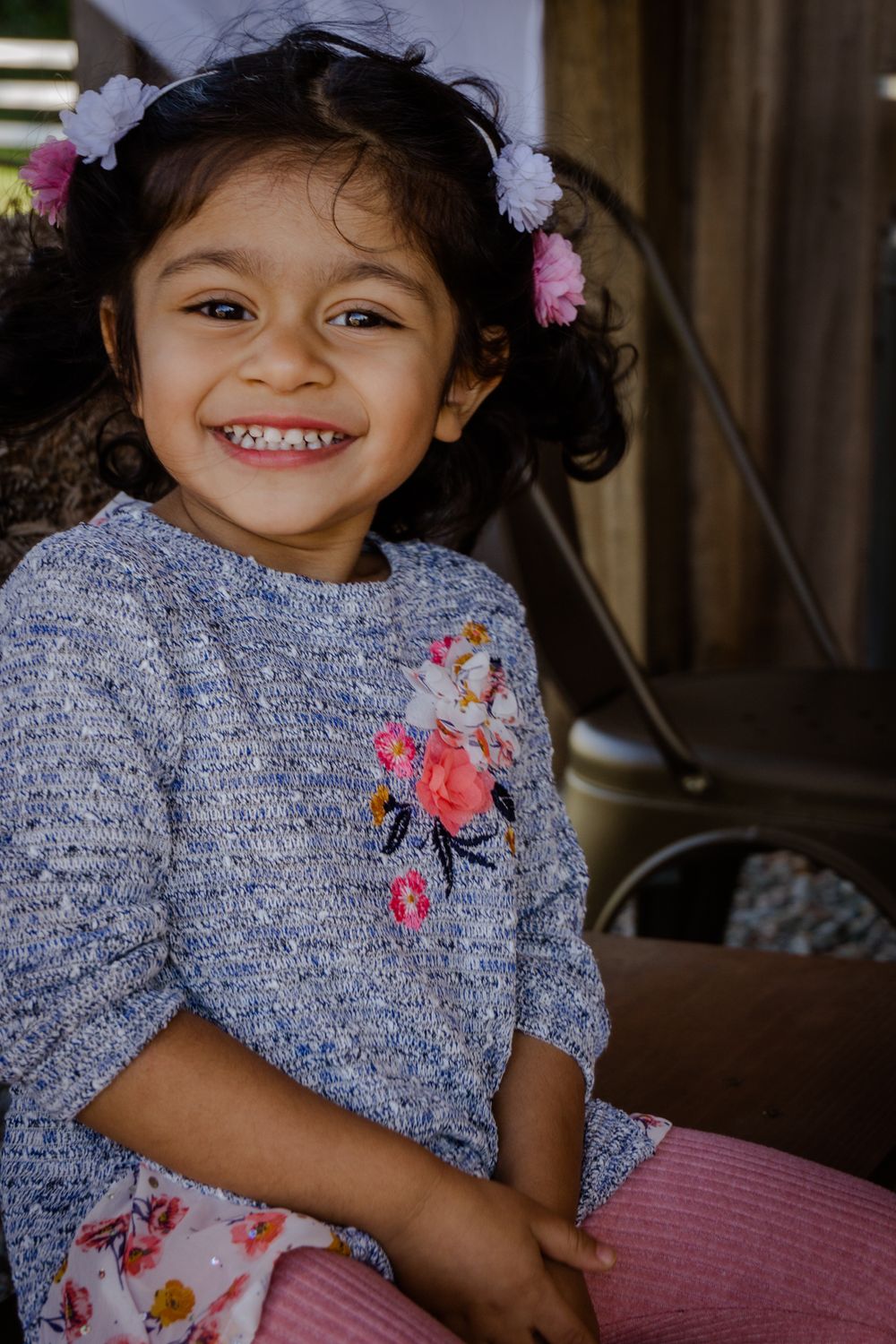 Smiling young girl with floral hair accessories wearing a gray knit sweater with embroidered flowers.