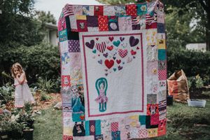 An art memory quilt showing a young dancer with hearts coming out of her hands is held up in a yard with a child playing