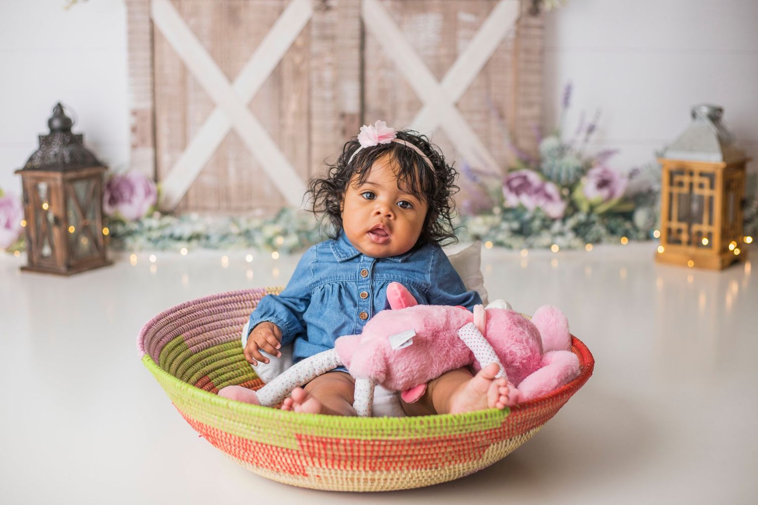 Baby in denim dress sitting in colorful woven basket with pink plush toy.