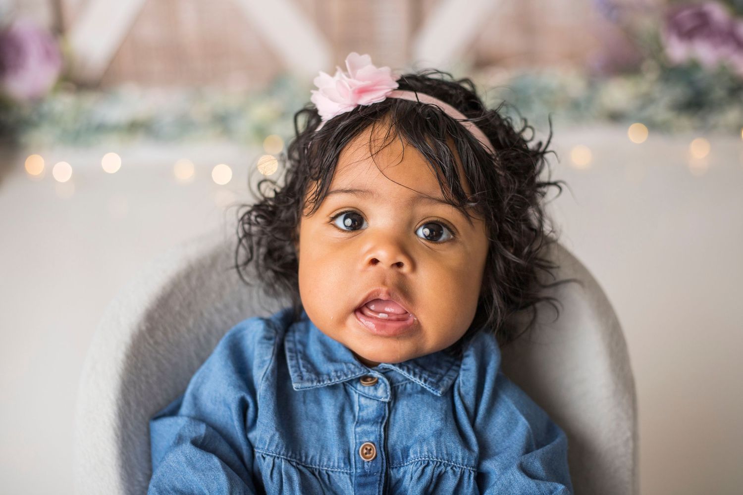 Baby in denim outfit with floral headband sitting in chair against rustic backdrop.