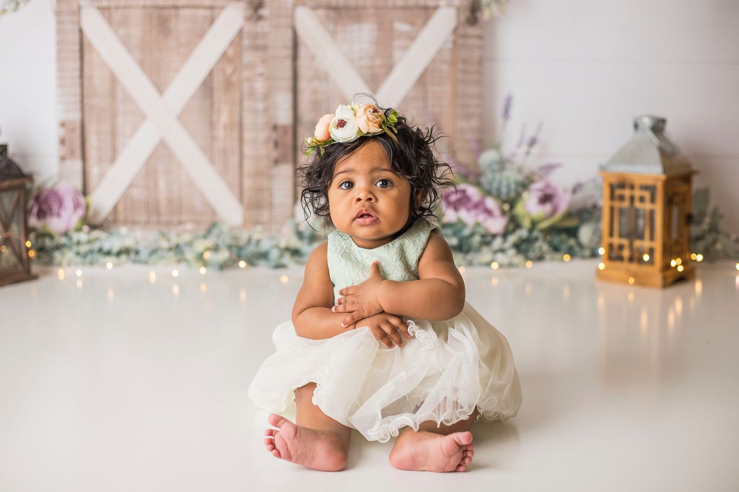 Baby in white dress with floral crown sits on studio floor with rustic barn door backdrop and lantern decor.
