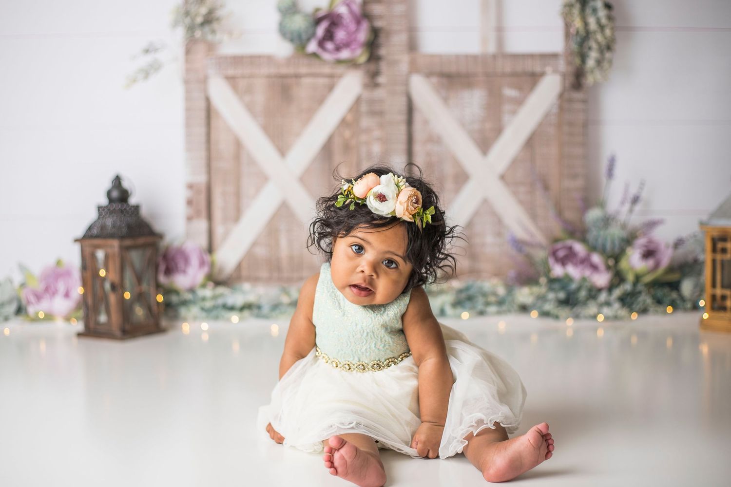 Baby wearing floral crown and white dress sits on floor with rustic barn door and floral backdrop.
