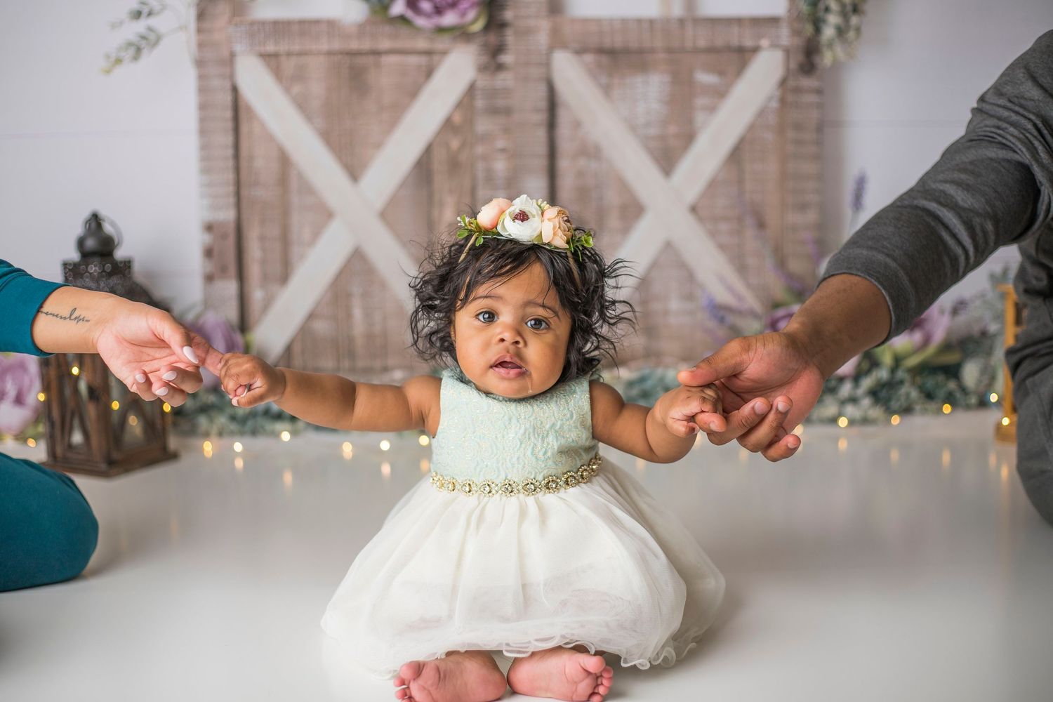 Baby in white dress holding parents' hands while sitting on studio floor with barn door backdrop.