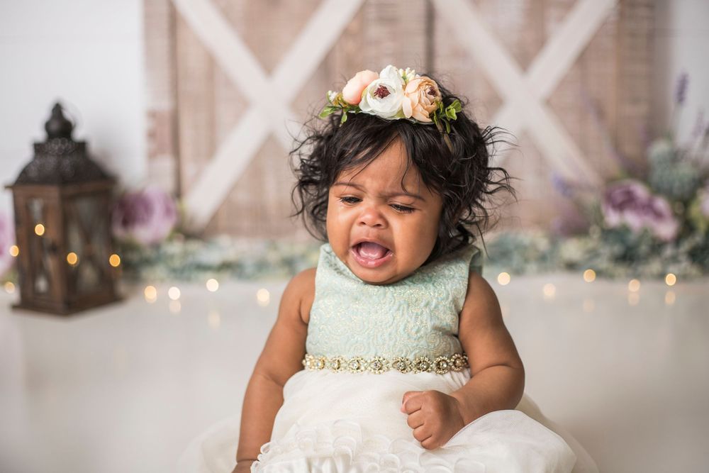 Baby wearing white dress and floral crown showing emotional expression in front of rustic backdrop.