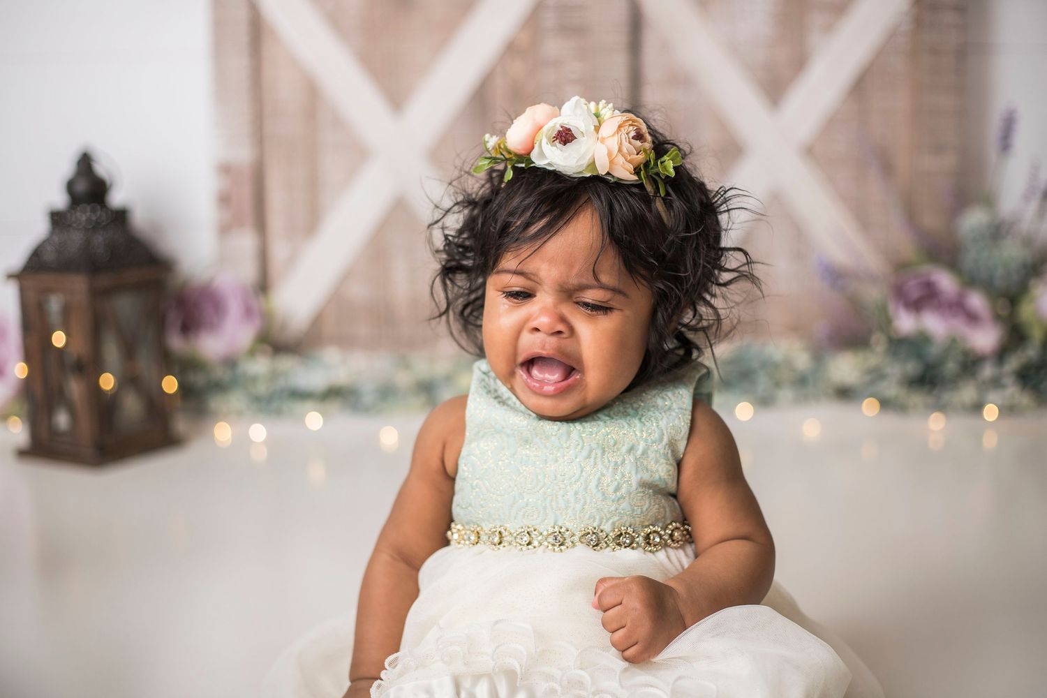 Baby wearing white dress and floral crown showing emotional expression in front of rustic backdrop.