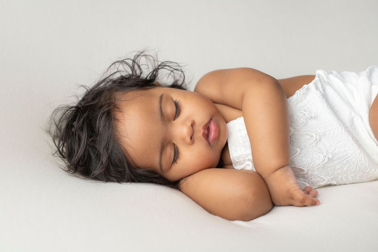 Peaceful sleeping baby in white outfit lying on light background.