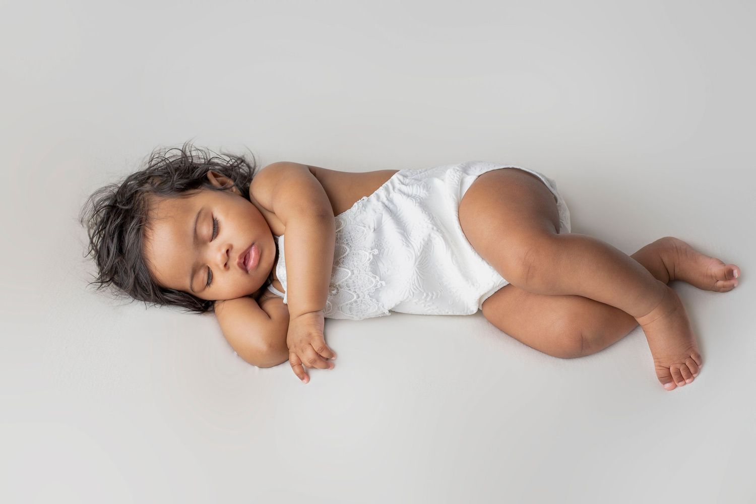 Baby in white outfit sleeping peacefully on light background in studio setting.