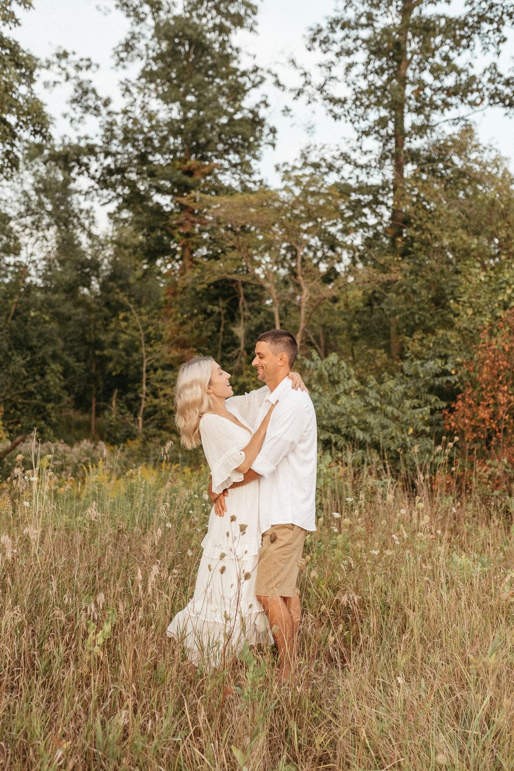 Couple embracing in tall grass field with autumn trees in background.