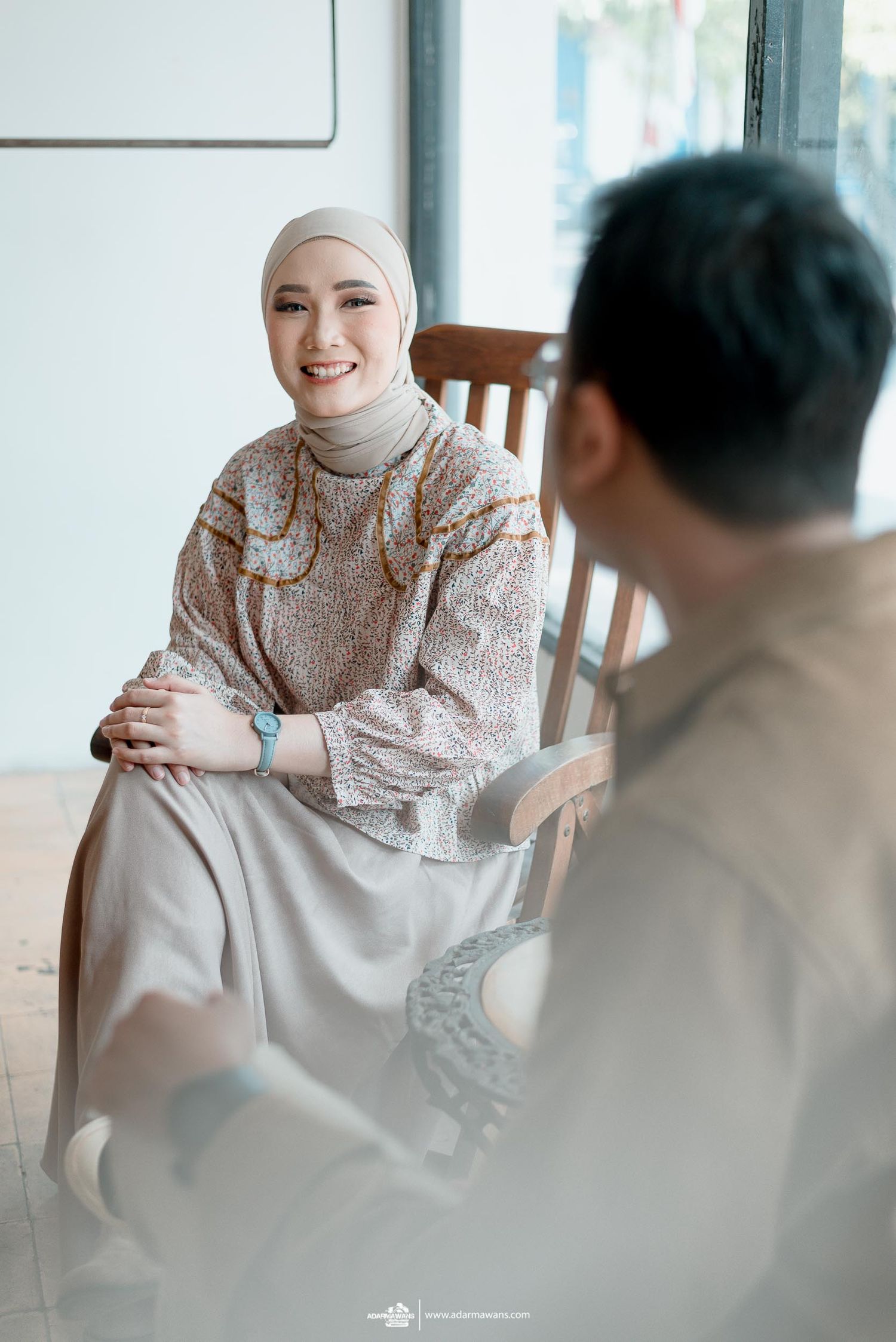 Person wearing a hijab and floral dress sits smiling during an intimate conversation at a cafe.