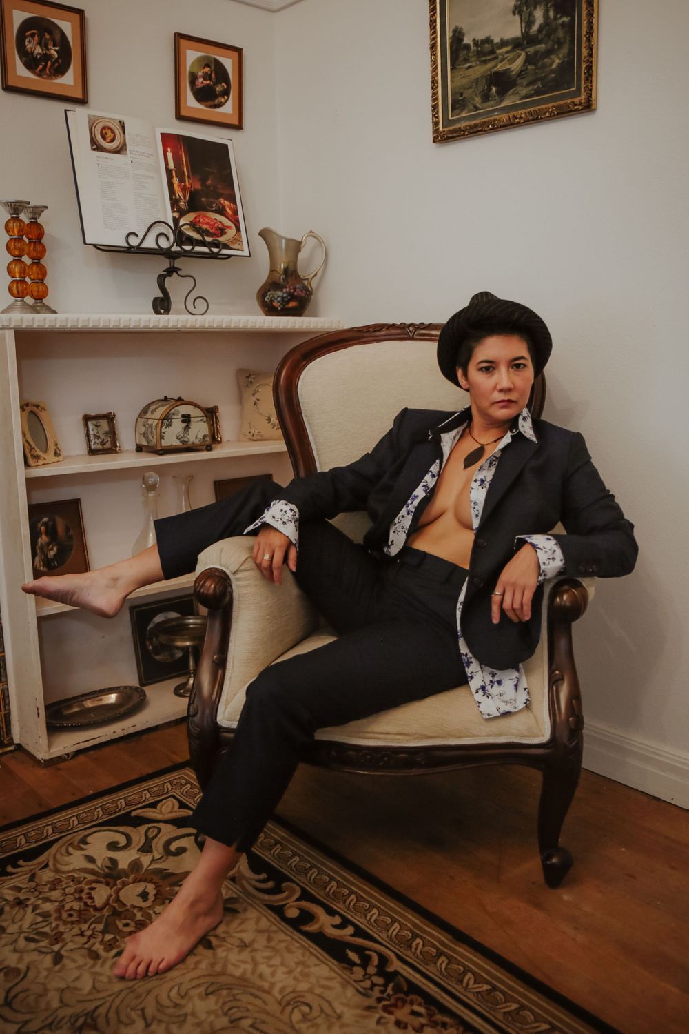 Person in dark blazer posing casually on vintage chair in room with decorative art and rugs.
