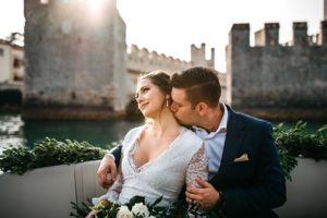 Bridal couple with the husband kissing the brides neck on a boat with the castle of Sirimione, Italy, in the background.