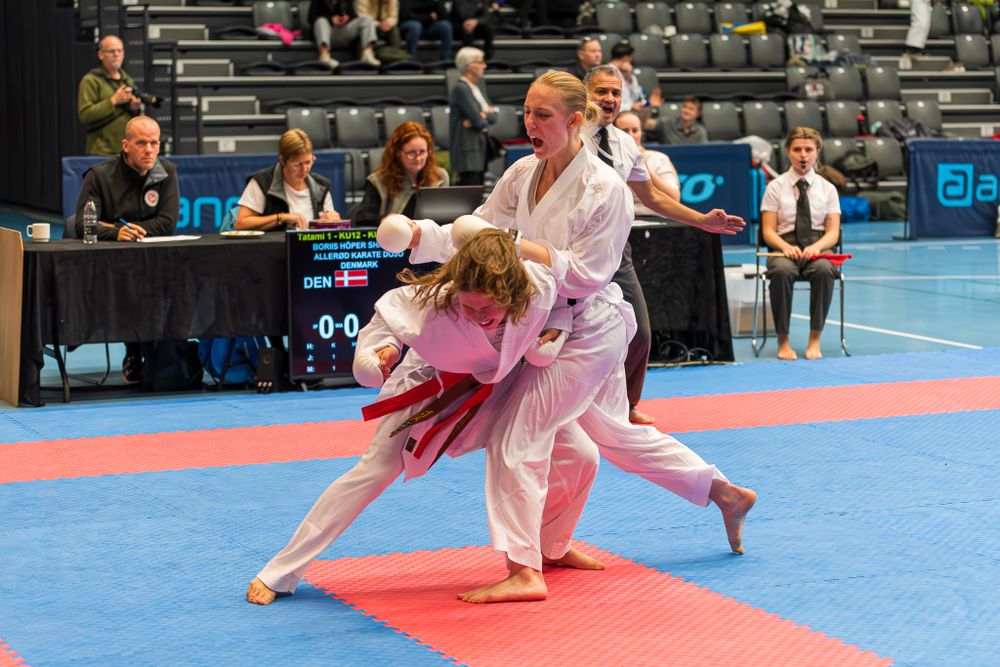 Female karate athletes grapple during competitive match on blue and red mat at indoor tournament.