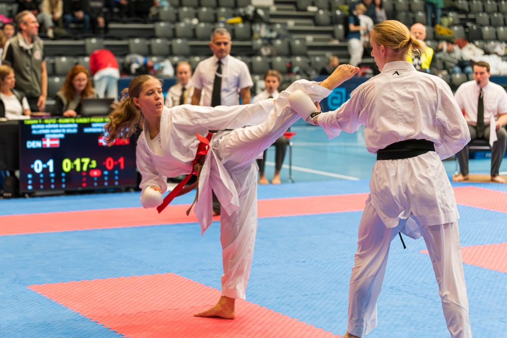 Female karate athlete executes high kick toward opponent during competitive tournament match.