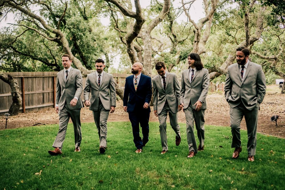 Groomsmen in matching gray suits jumping playfully on green lawn.