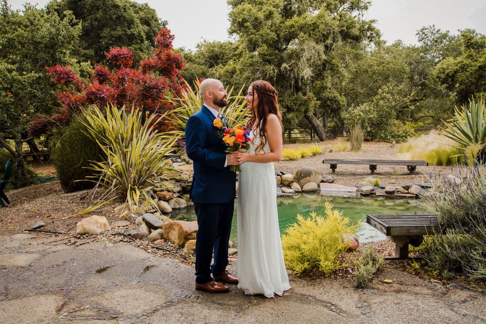 Couple sharing intimate moment in garden setting with pond and colorful plants in background.