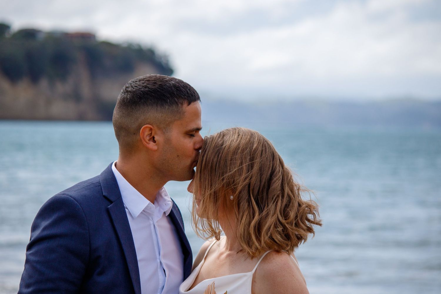 Newlywed couple sharing a romantic kiss by the ocean with coastal cliffs in background.