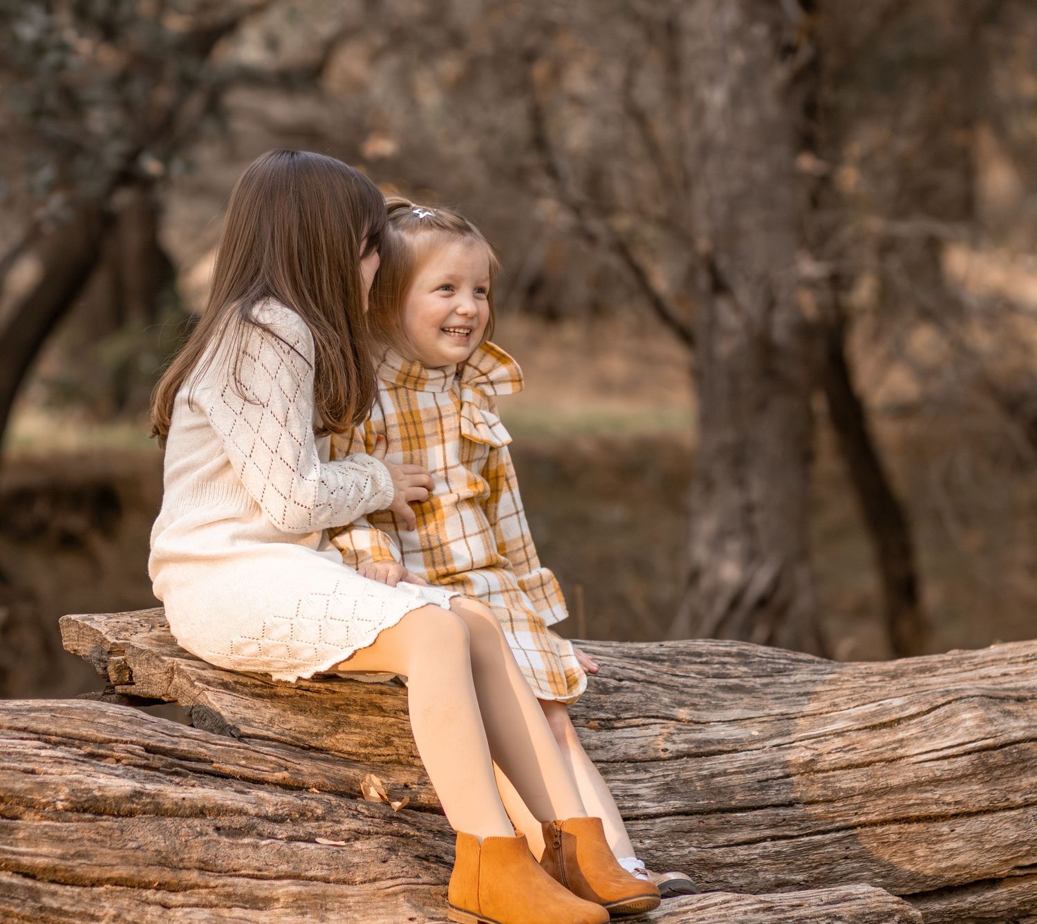 Two young sisters sitting on a log in autumn woods, older girl hugging smiling younger sister.
