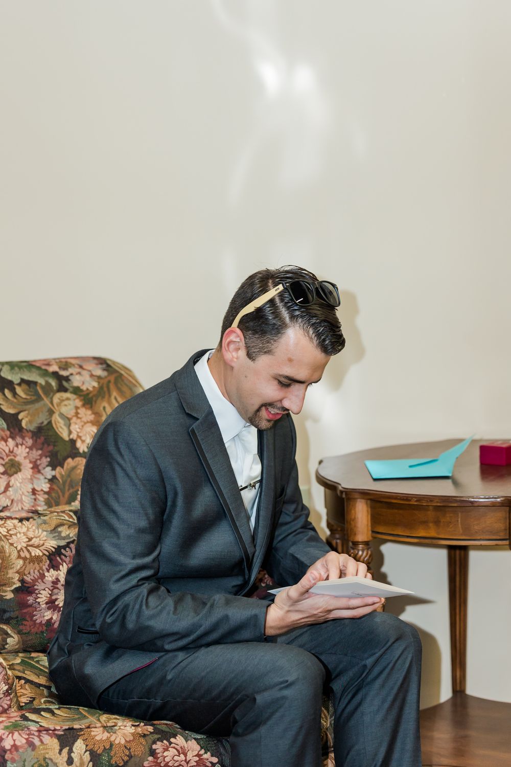 Groom in dark suit sits in floral chair while writing in a journal during wedding preparations.