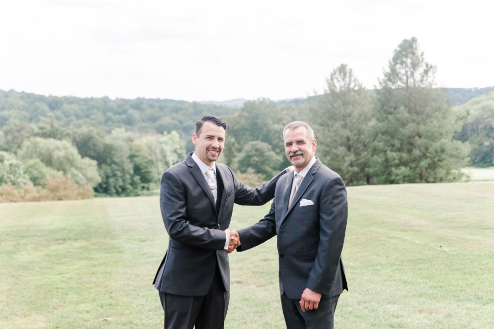Two men in suits shake hands while standing on a grassy hillside.