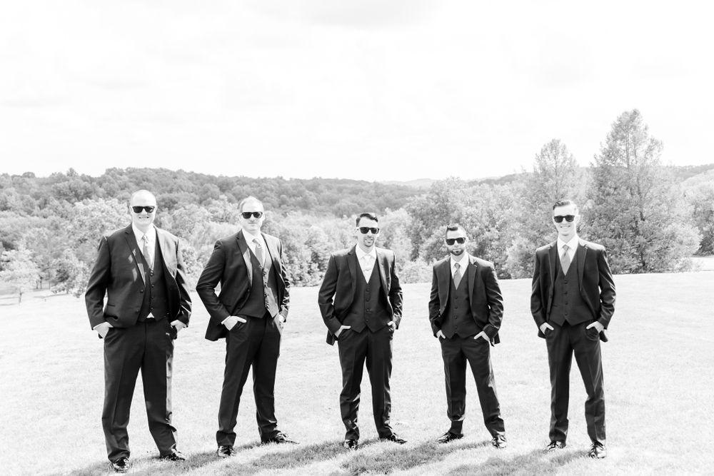 Five groomsmen in black suits and sunglasses pose together on a scenic hillside.