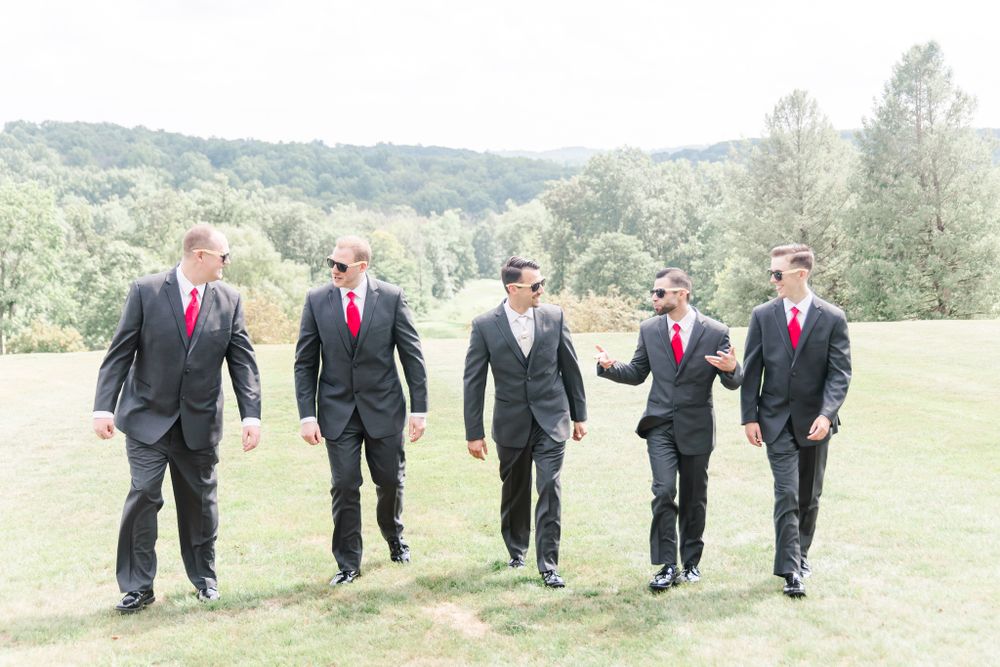 Five groomsmen in dark suits and red ties walking together across a scenic hillside.
