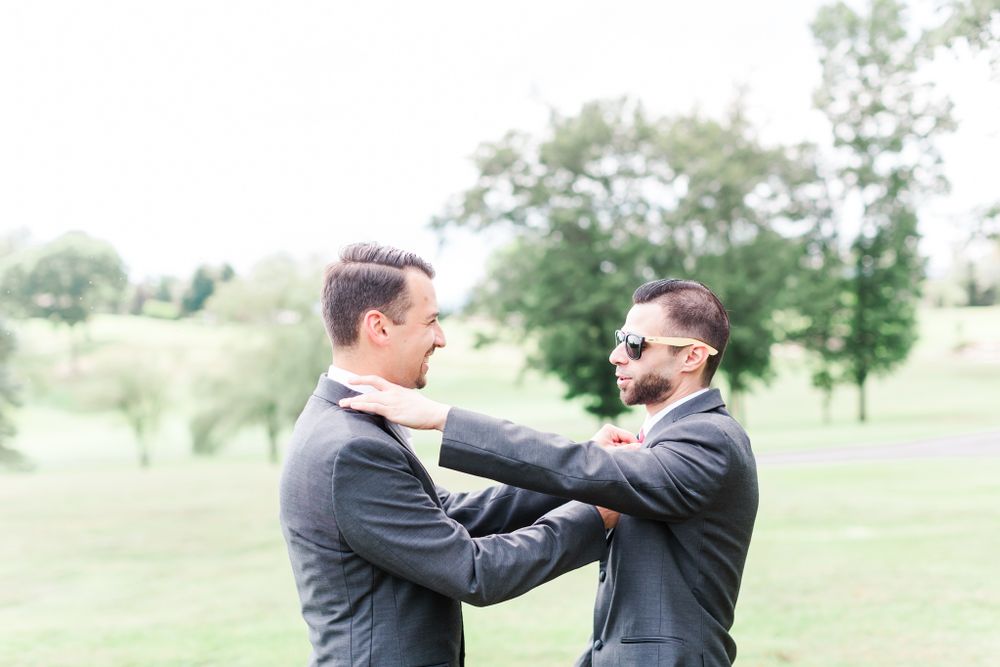 Two groomsmen in suits share a playful moment adjusting each other's attire outdoors.