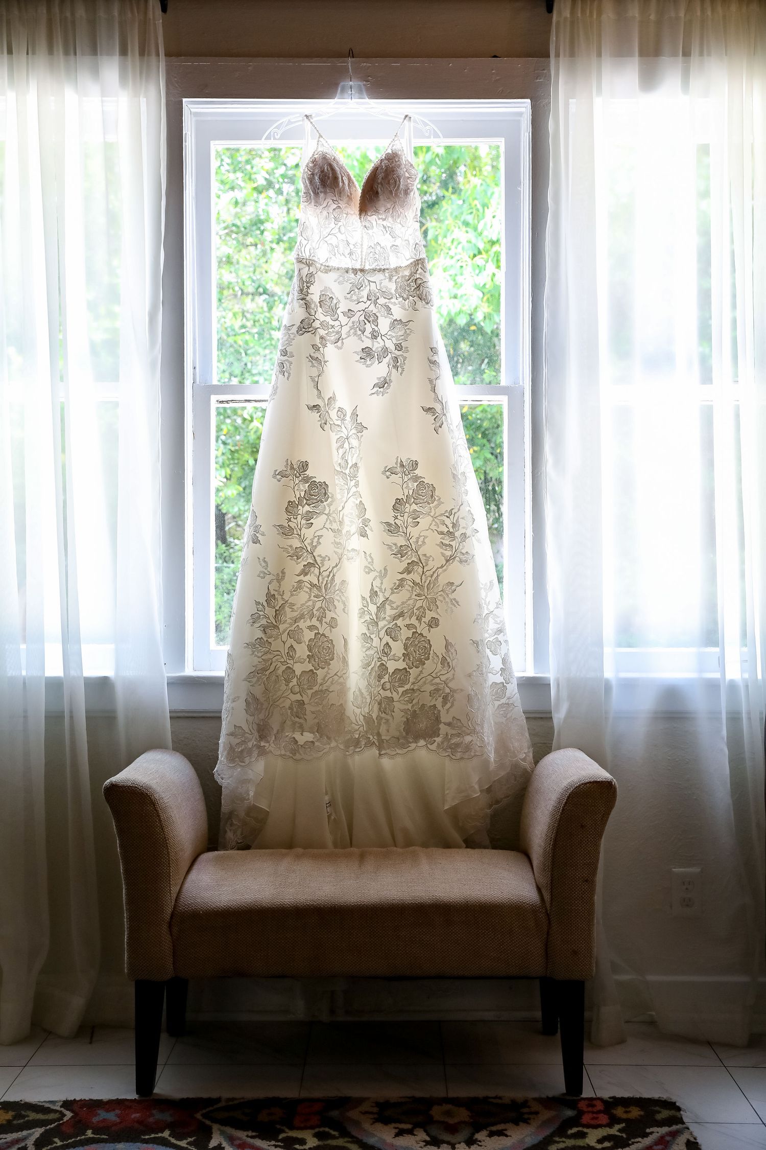 An elegant white wedding dress with lace details hangs in front of a bright window.