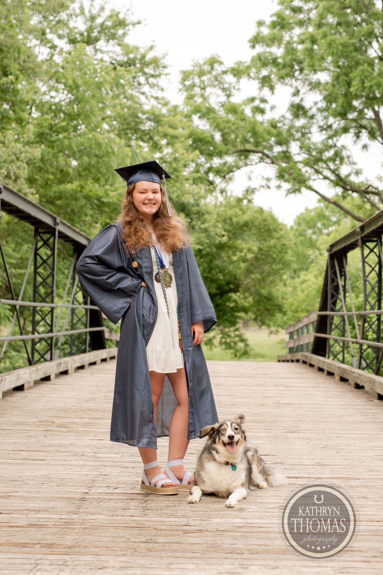 Graduate in academic regalia poses with dog on wooden bridge for commencement photos.