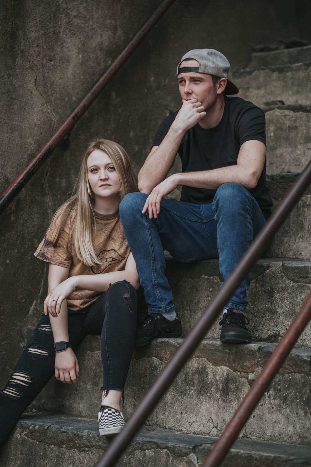 Relaxed lifestyle portrait of pair sitting on worn concrete steps against industrial backdrop.