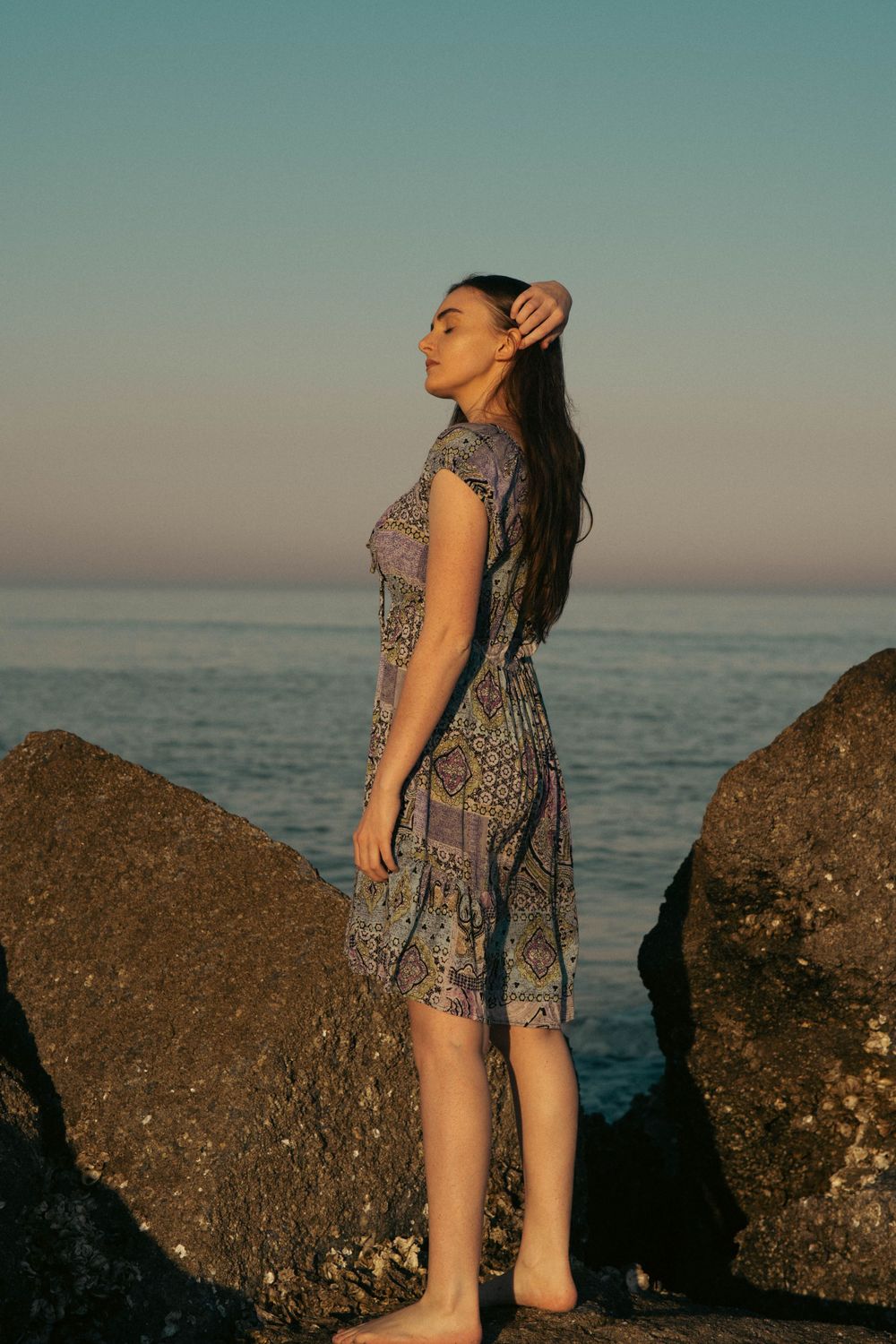 A peaceful coastal portrait showing someone standing on rocks by the ocean at sunset.