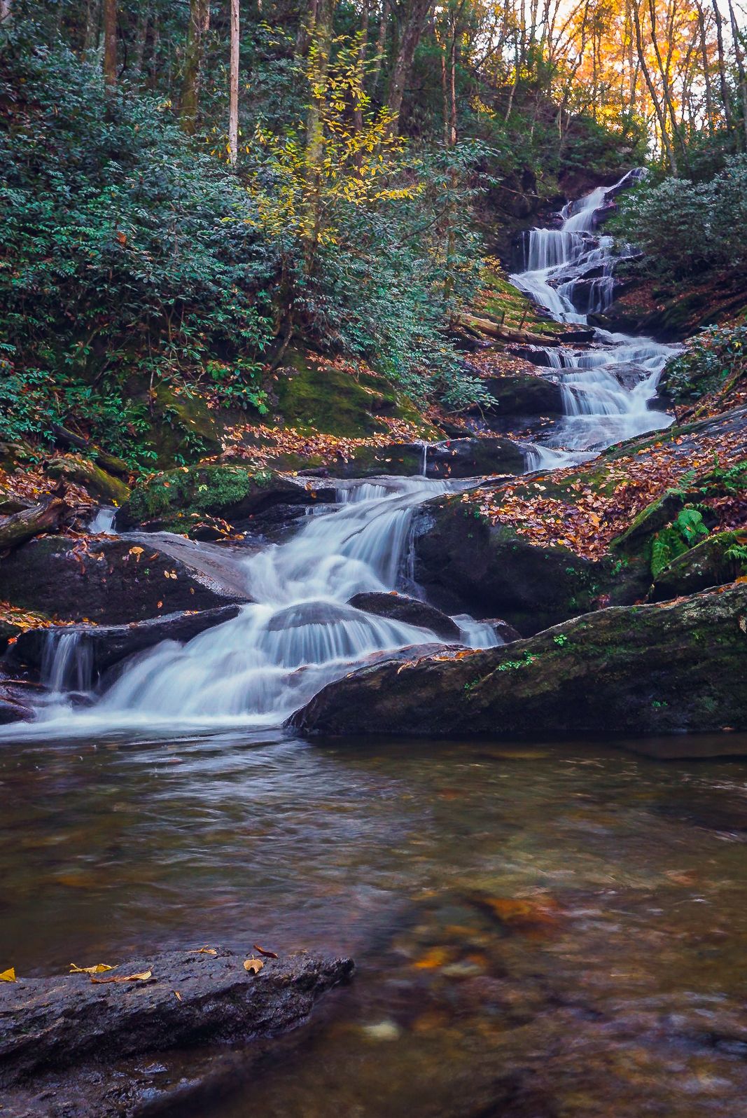 A long cascade winds through boulders surrounded by gold, green and orange leaves in this long exposure photograph.