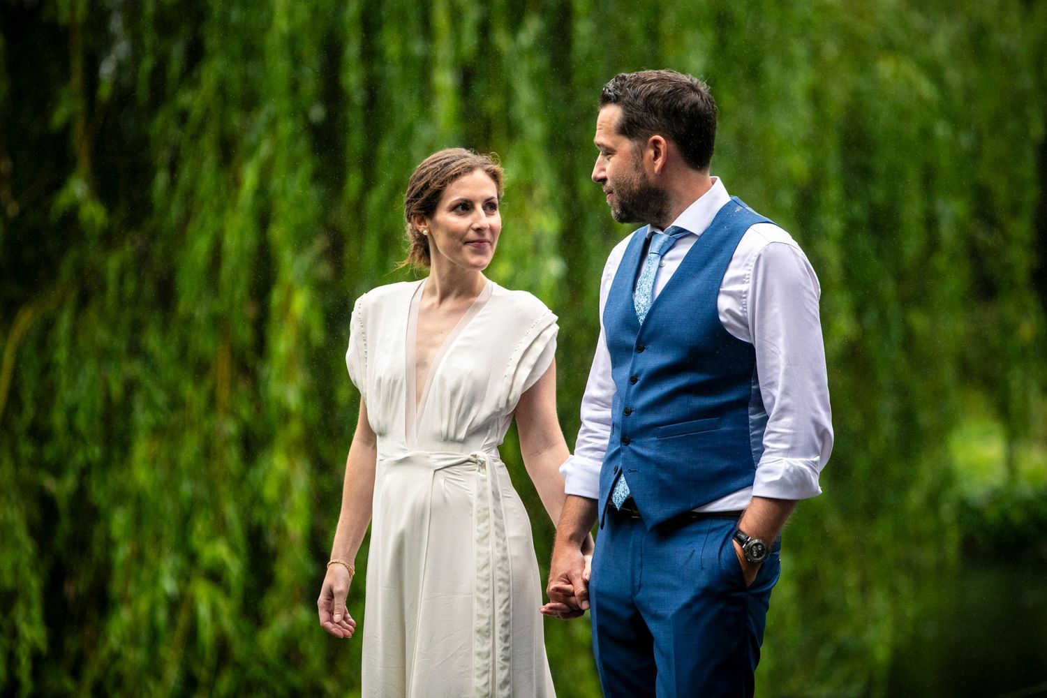 Couple in wedding attire poses together against a backdrop of weeping willow trees.