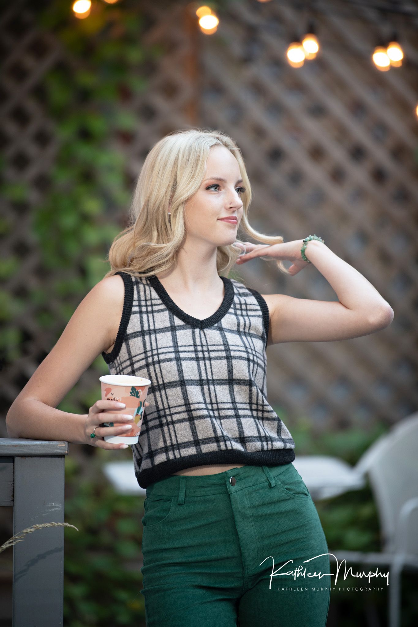 A fashion portrait of someone in a plaid crop top and green pants holding a coffee cup against a latticed background.