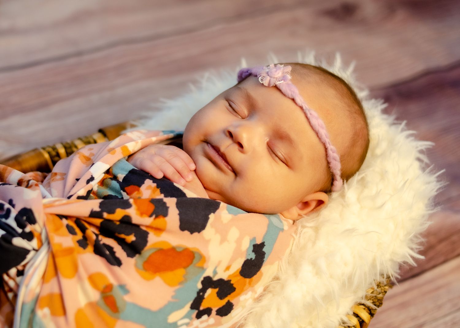Sleeping baby in basket with fur blanket wearing a purple headband wrapped in multi-colored blanket while getting gentle newborn photos