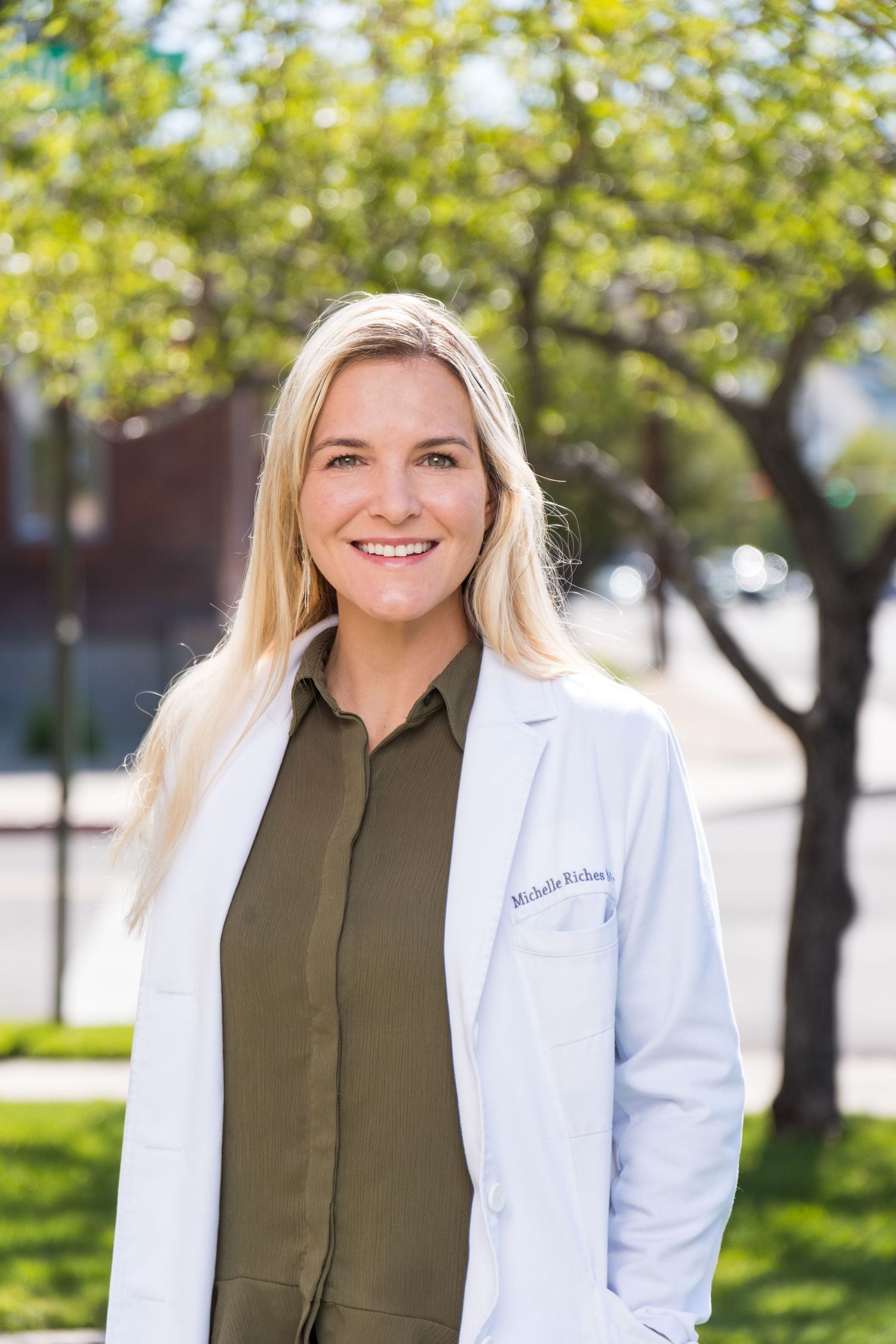Medical professional in white lab coat and olive top smiling outdoors on sunny day with trees in background.