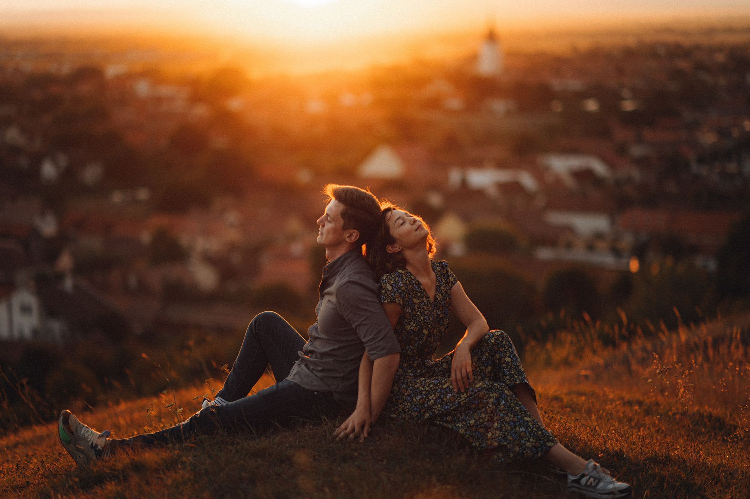 Two figures sit together watching a golden sunset over a cityscape in warm atmospheric lighting.