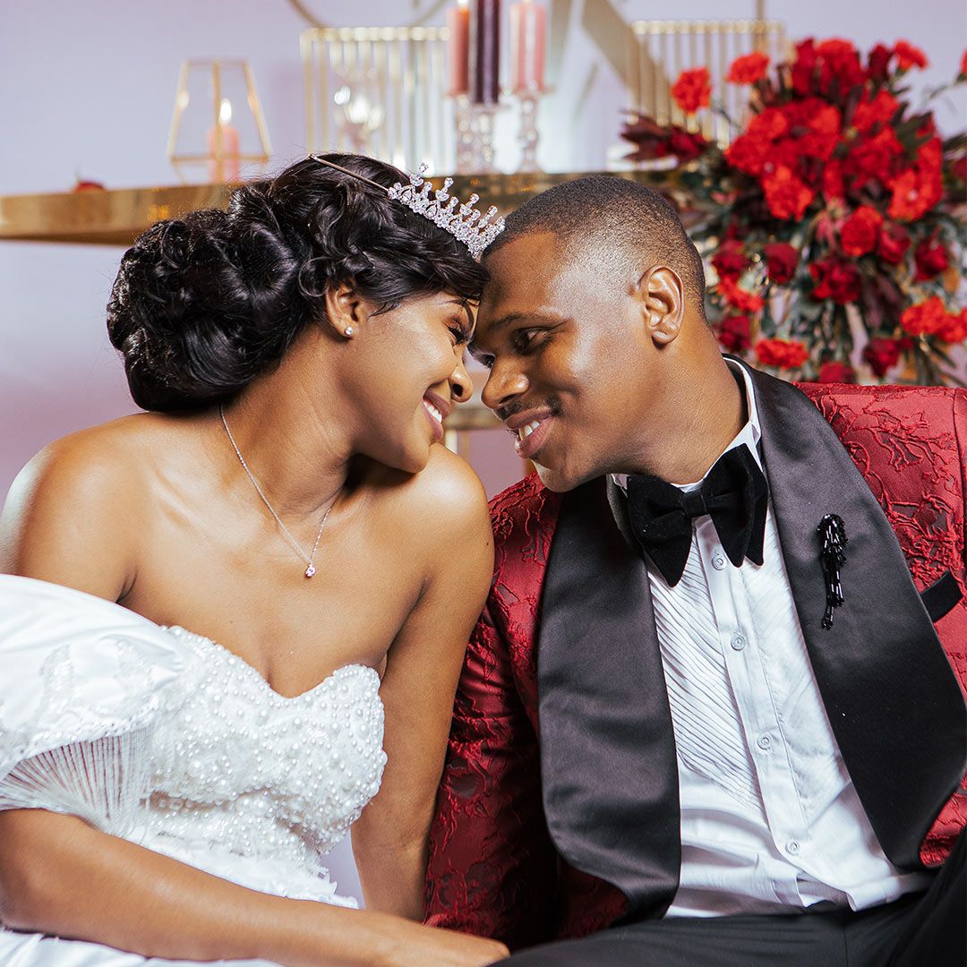 Romantic wedding portrait with red floral decor and elegant attire including tiara and bow tie.
