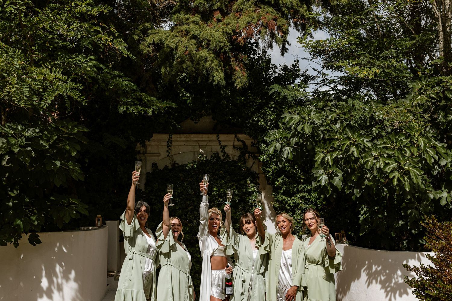 Wedding party wearing sage green dresses raises their arms in celebration against a sunlit garden wall at Quinta da Bichinha