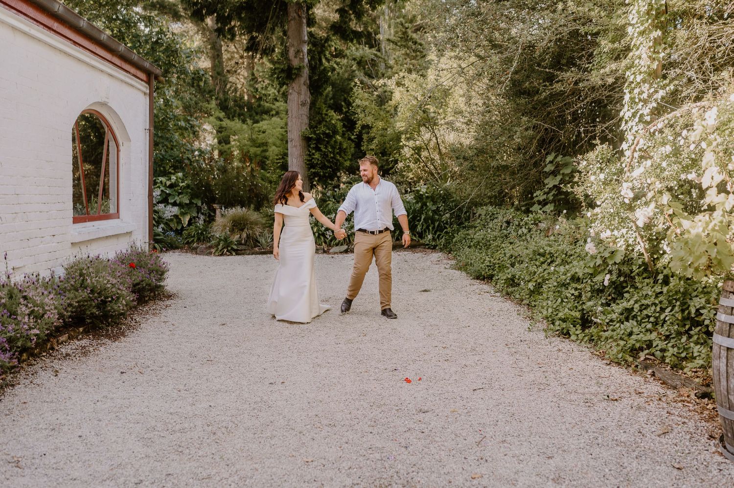 A couple strolls together on a gravel path next to a white building with an arched window.