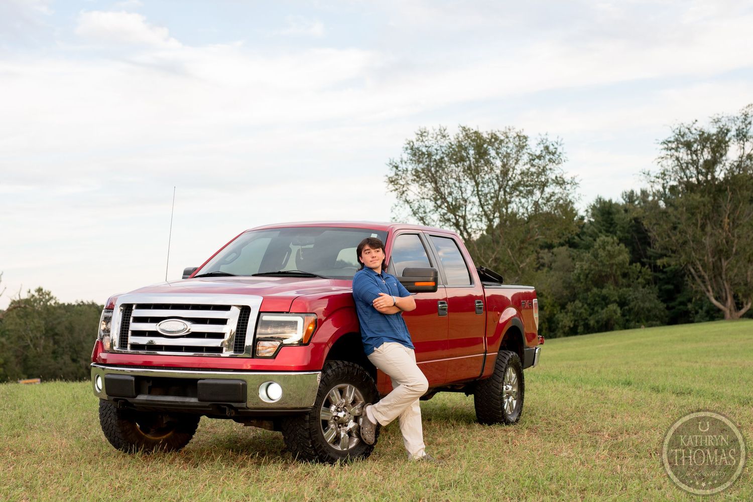 Someone poses with a lifted red Ford F150 pickup truck in a scenic field setting.