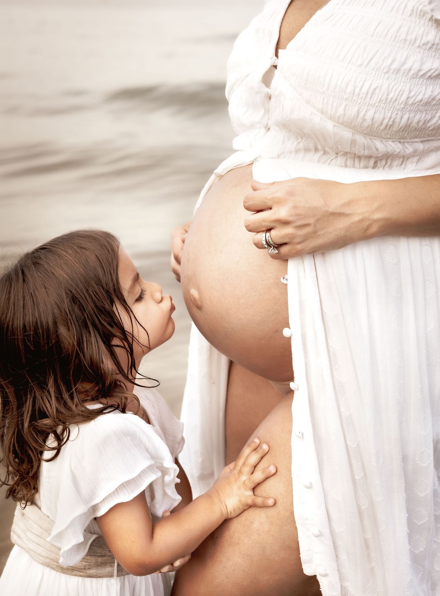 Intimate maternity photo with child touching pregnant belly in white dress.