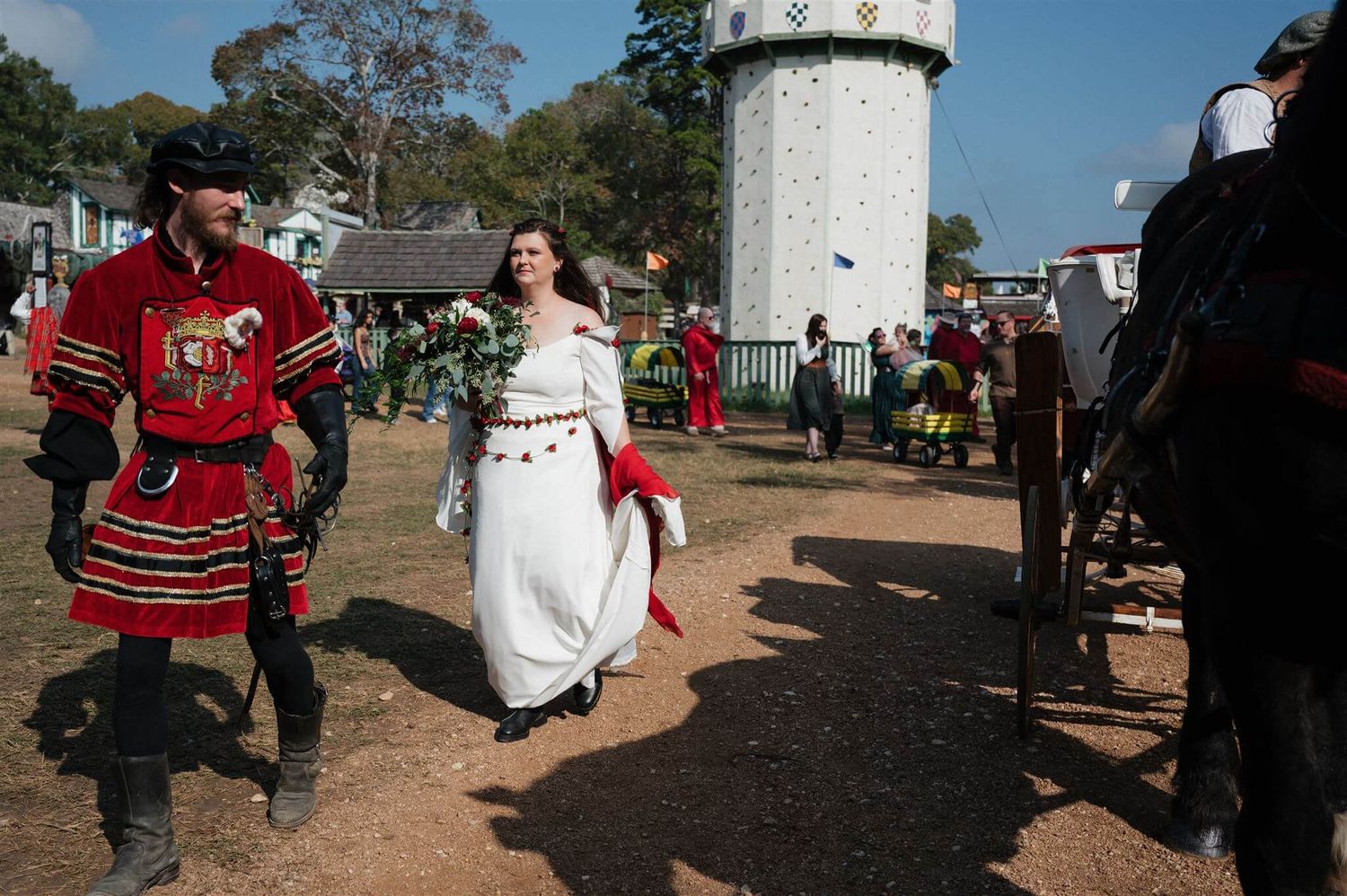 A person in medieval red attire walks alongside another in a white dress at an outdoor festival.