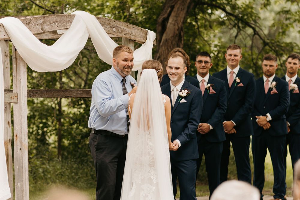 Groom smiling at altar with groomsmen in navy suits lined up beside rustic wooden wedding arbor.
