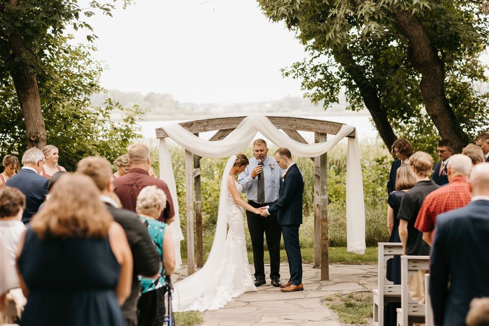 Wedding ceremony couple holding hands before officiant at rustic wooden arbor with white fabric draping.