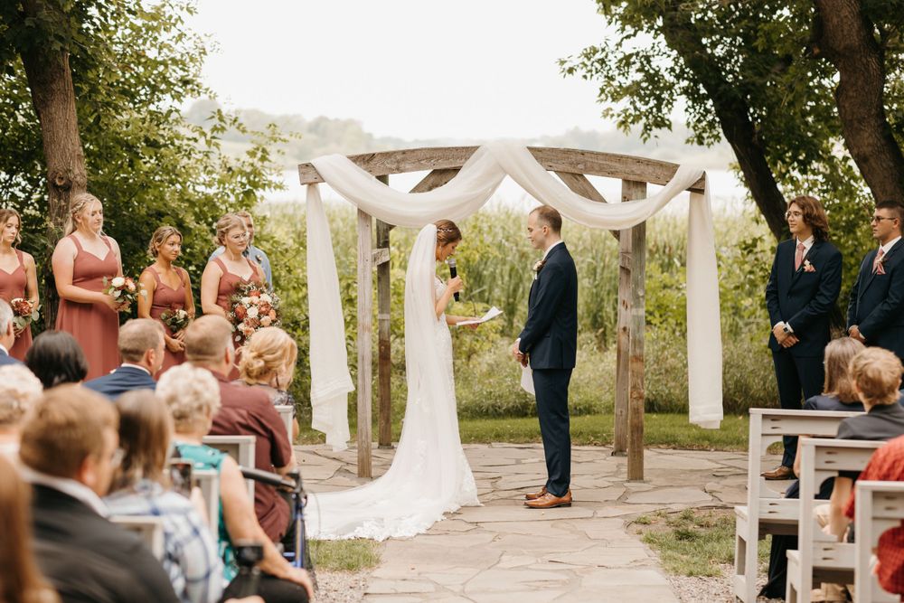 Outdoor wedding ceremony with bride and groom at wooden arbor, guests seated on benches among trees.