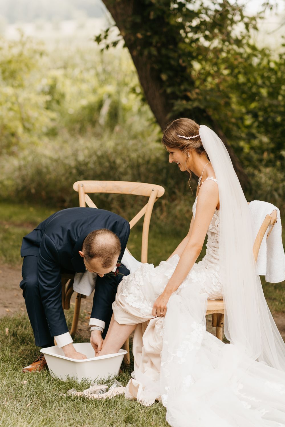 Groom washing bride's feet in a white basin during an outdoor wedding ceremony unity ritual.