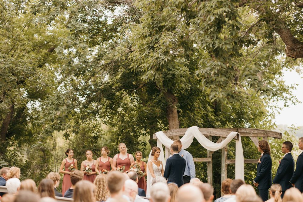 Outdoor wedding ceremony under large trees with bridesmaids in dusty rose dresses standing at a rustic wooden arch.